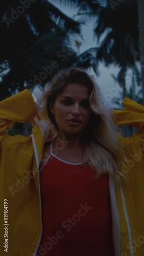 A beautiful girl in a yellow raincoat is walking along the road among the palm trees. A sexy woman in a coat is flirting with the camera. A blonde female enjoys the tropical island of Bali. photo