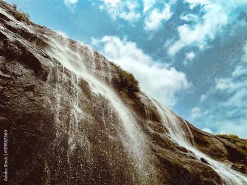 Splashing waterfall on rock mountain with clouds sky in background, low angle view of splashing droplets of stram fresh water from the hill photo