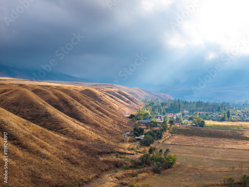 View of autumn landcape with mountains, village and field. Large clouds in the sky, the sun's rays shine through the clouds. Yellow fields near the hills. Vivid natural scenery photo.