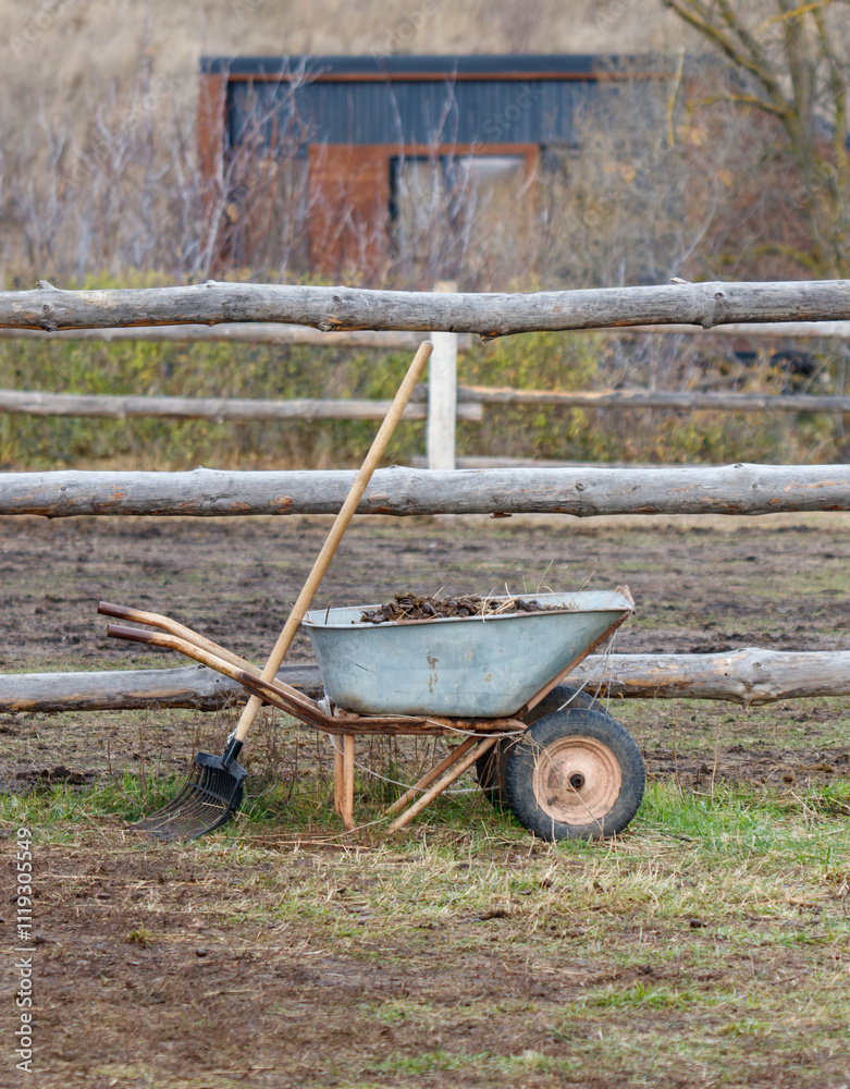 A wheelbarrow with a rake and a wooden fence in the background A wheelbarrow with a rake and a wooden fence in the background