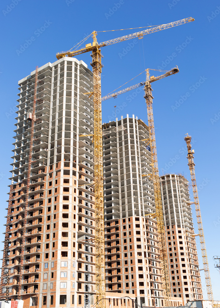 Three modern high-rise panel new building under construction and three tower cranes against the blue sky, vertical photo. Three modern high-rise panel new building under construction and three tower cranes against the blue sky, vertical photo.