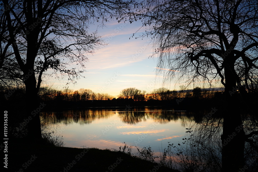 An idyllic panorama at the Black Lake (Schwarzer See) in Garbsen in winter. Reflections of the clouds in the water after sunset. Frame of leafless trees in frosty night. Garbsen, Lower Saxony, Germany An idyllic panorama at the Black Lake (Schwarzer See) in Garbsen in winter. Reflections of the clouds in the water after sunset. Frame of leafless trees in frosty night. Garbsen, Lower Saxony, Germany
