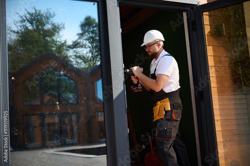 Construction worker installing door in a new house construction site. Man in working attire install front door. Builder in uniform with screwdriver adjusting front glass door in new beautiful house.