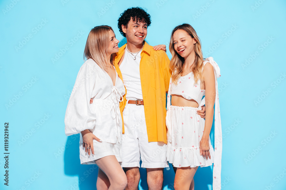 Group of young three stylish friends posing in studio. Fashion man and two cute female dressed in casual summer clothes. Smiling models having fun. Cheerful women and guy near blue wall