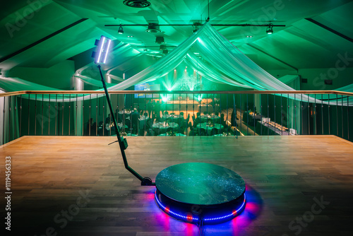  Elevated view of a green-lit event venue featuring a 360 video booth platform, draped ceilings, banquet tables, and elegant lighting decor. photo