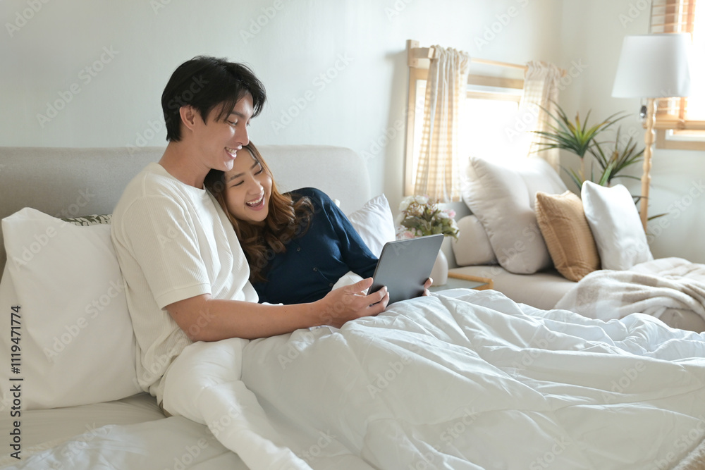A man and woman are lying in bed together, with the man holding a tablet. A man and woman are lying in bed together, with the man holding a tablet.