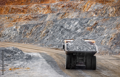 Heavy dump truck transporting gold ore load in open pit mine landscape, back view.