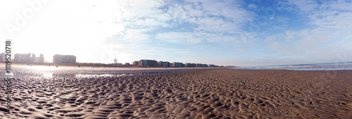 A panoramic view of a vast sandy beach with a city skyline under a bright blue sky. The tranquil scene evokes peacefulness and escape, perfect for a background or travel concept.
