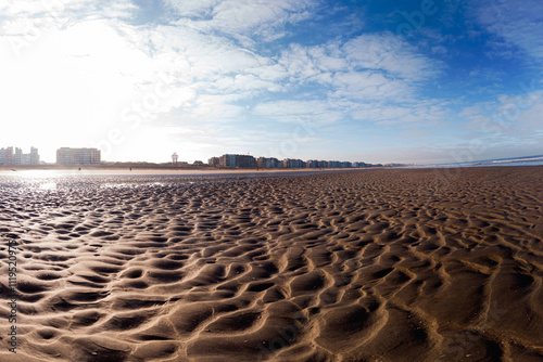 A wide sandy beach with intricate patterns stretches towards a distant city skyline under a vibrant blue sky. Serene and inviting, the scene captures the tranquility of a coastal landscape.