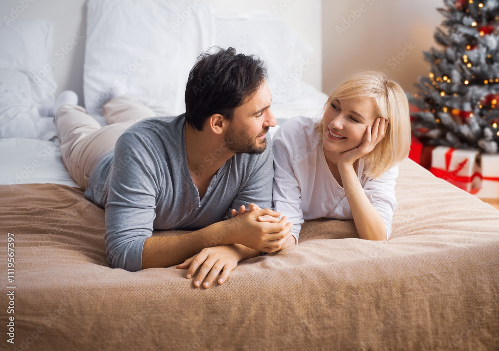 Image of smiling couple, husband and wife looking at each other on bed, Christmas tree, gifts, lights home background. Woman, man in love, people, happy family, winter holidays celebration - concept Image of smiling couple, husband and wife looking at each other on bed, Christmas tree, gifts, lights home background. Woman, man in love, people, happy family, winter holidays celebration - concept