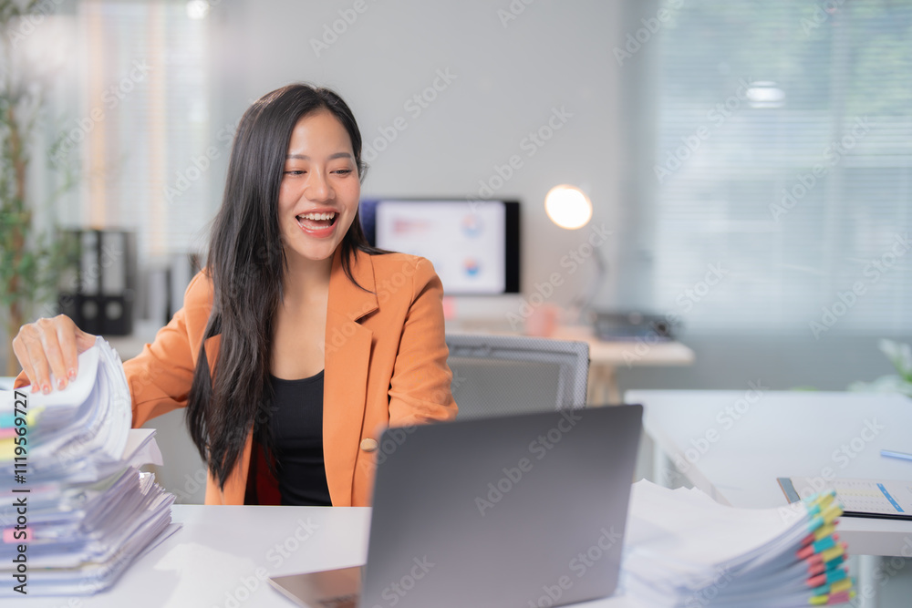 Asian businesswoman is smiling and holding documents while having a video conference on her laptop in a bright office, she is wearing an orange blazer and a black shirt Asian businesswoman is smiling and holding documents while having a video conference on her laptop in a bright office, she is wearing an orange blazer and a black shirt