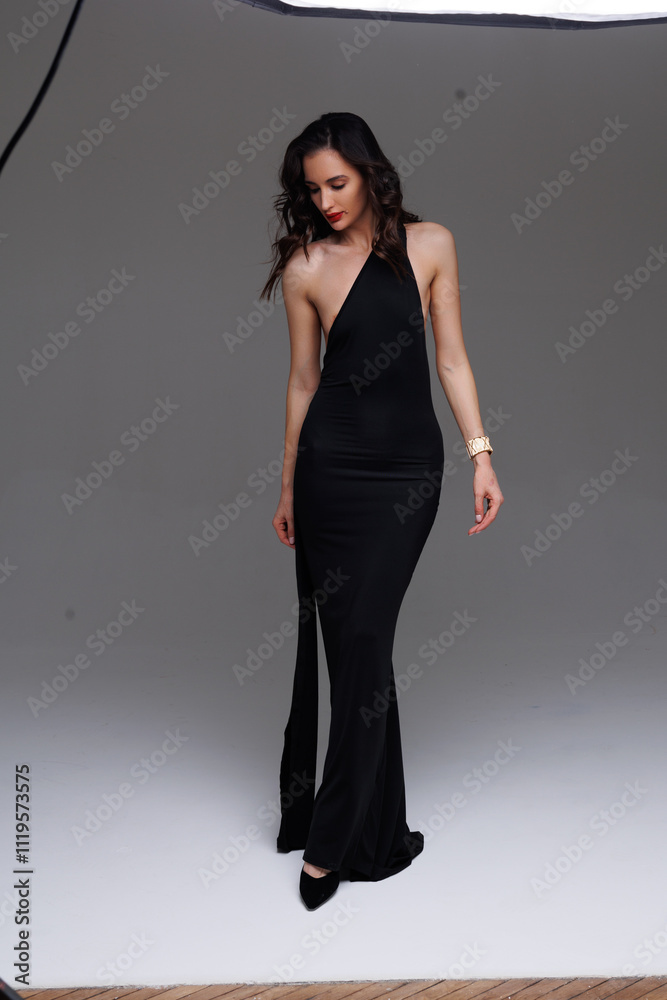 A gorgeous tall slender woman model in a black dress with heels stands at grey background. Thin gorgeous woman. A brunette girl with a good figure on heels smiles