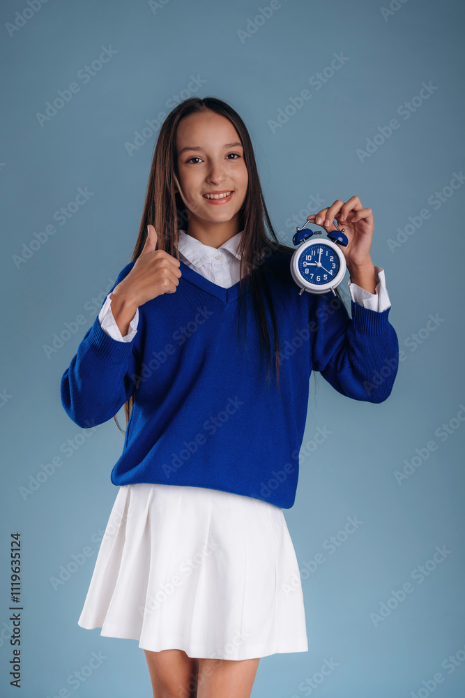 Fototapeta premium With blue clock in hand. Young schoolgirl in uniform is against background