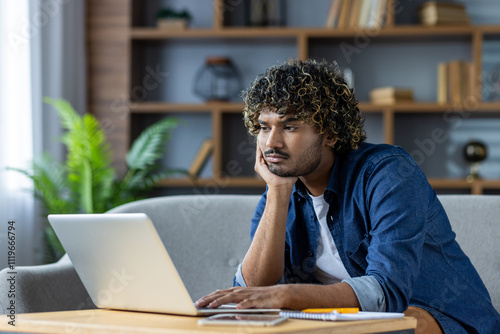 A young man with curly hair appears bored while using a laptop at home. He rests his head in his hand, illustrating a common work-from-home experience. This image captures feelings of monotony