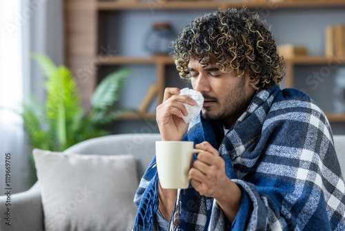 A man wrapped in a plaid blanket, battling a cold indoors, holds a cup and tissue. He appears to be sneezing, suggesting symptoms typical of flu or allergies.