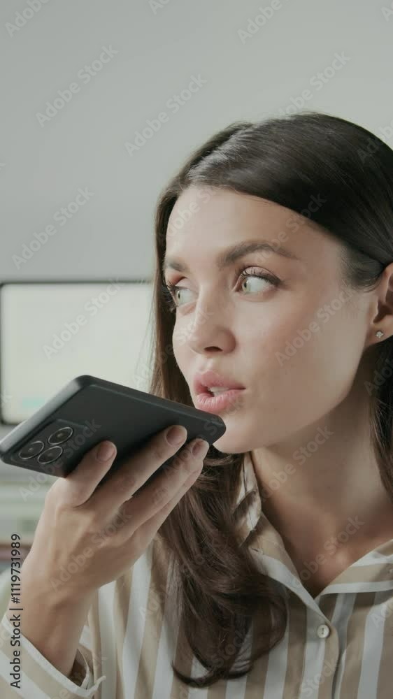 Waist up of young Caucasian businesswoman in striped shirt texting and recording messages on her smartphone while working at desk in bright, modern office, vertical shot Waist up of young Caucasian businesswoman in striped shirt texting and recording messages on her smartphone while working at desk in bright, modern office, vertical shot