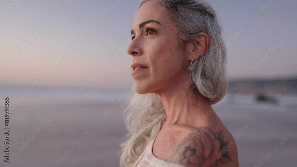 A thoughtful mature woman with gray hair and a tattoo reflects on life's journey while enjoying a peaceful sunset at the beach.