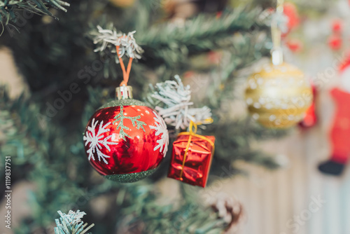 Brightly colored ornaments hang on s tree, featuring red ball with snowflake designs and shiny gold ornament alongside small red gift box photo