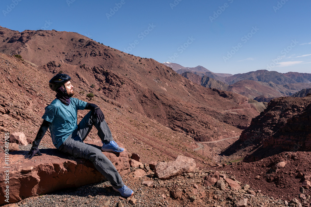 Serenity in the Atlas. Cyclist Resting on a Rock, Immersed in Peace and the Majestic Mountain Landscape Serenity in the Atlas. Cyclist Resting on a Rock, Immersed in Peace and the Majestic Mountain Landscape