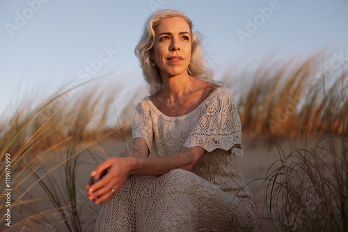 Canvas Print A mature woman with gray hair sits calmly on a sandy beach, basking in the warm golden hour sunlight