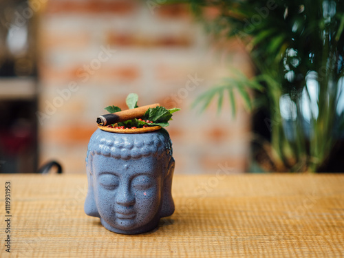 Buddha-shaped ceramic mug cocktail with intricate details, garnished with a cinnamon stick and mint leaves, photographed against a soft-focus brick wall and greenery background, creamy bokeh.