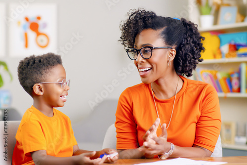 African American woman speech therapist and a young boy engage in a fun and interactive conversation, both smiling and wearing bright orange outfits, sitting in a well-lit and colorful therapy room.