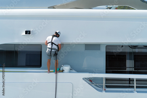 Male Caucasian Deckhand working on the side of a white modern superyacht with harness and safety helmet while doing a washdown
