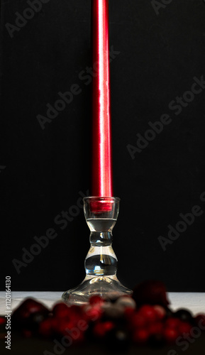 Red candle on glass stand in focus, foreground of red fruits out of focus, black background. Concept of love and valentine's day