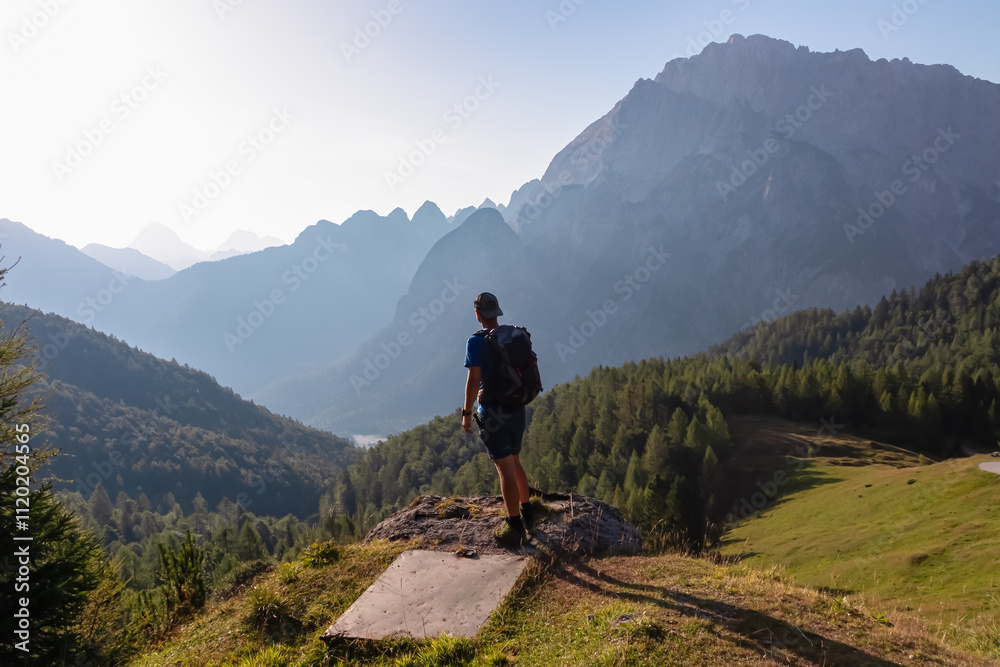 Hiker man with backpack looking at majestic mountains peaks Julian Alps in Somdogna, Friuli Venezia Giulia, Italy. Alpine pastures and slopes covered in coniferous forest. Wanderlust in Italian Alps Hiker man with backpack looking at majestic mountains peaks Julian Alps in Somdogna, Friuli Venezia Giulia, Italy. Alpine pastures and slopes covered in coniferous forest. Wanderlust in Italian Alps