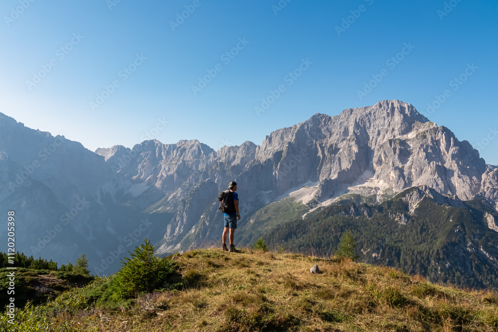 Obraz premium Active hiker man on alpine meadow with scenic view of rugged mountain peak Jof di Montasio. Majestic ridges of Julian Alps in Somdogna, Friuli Venezia Giulia, Italy. Wanderlust Italian Alps in summer