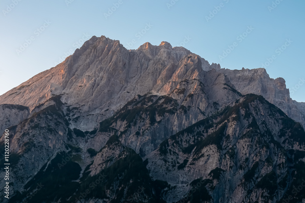 Naklejka premium Scenic view of stunning mountain peak Jof Fuart bathed in soft glow of twilight. Massive rugged ridges of majestic Julian Alps seen from Val Saisera, Valbruna, Friuli Venezia Giulia, Italy. Sunrise