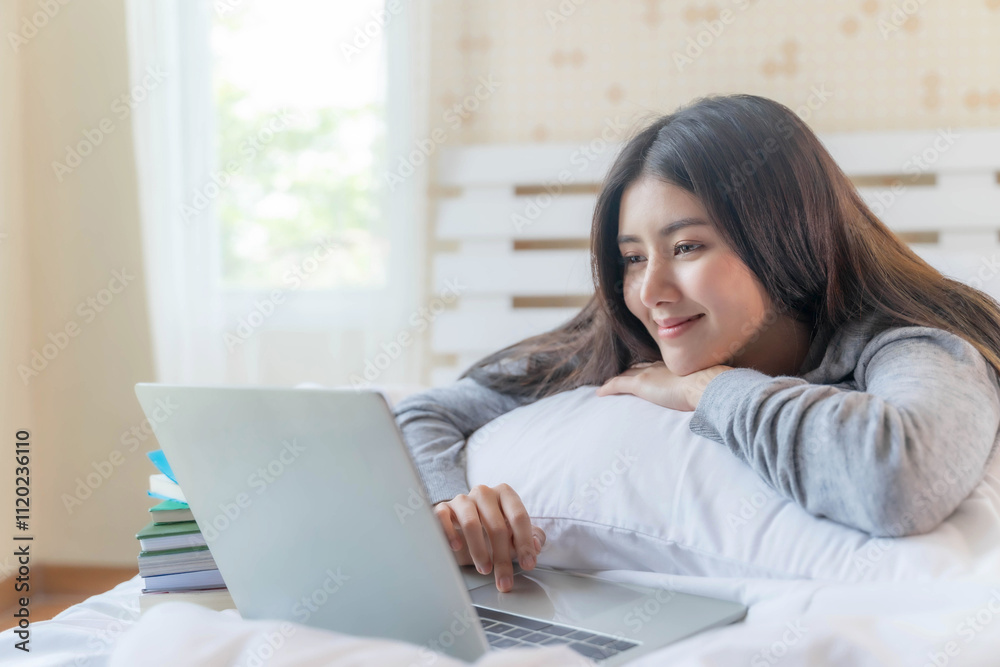 Relaxed young Asian woman lying on a bed, using a laptop at home, surrounded by books, enjoying a peaceful studying or working in a cozy bedroom environment Relaxed young Asian woman lying on a bed, using a laptop at home, surrounded by books, enjoying a peaceful studying or working in a cozy bedroom environment