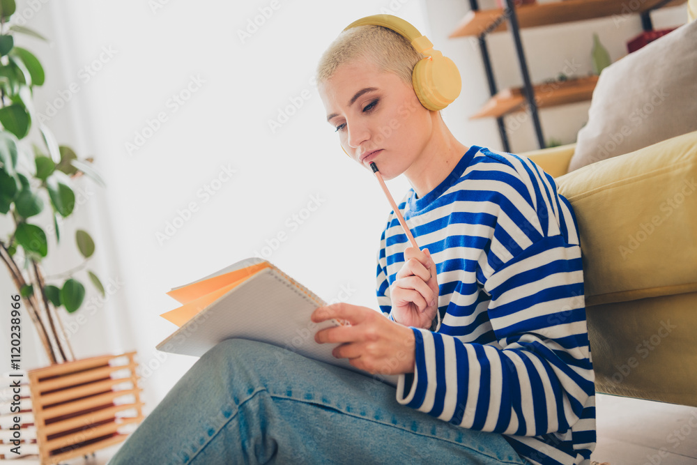 Young woman in striped pullover enjoying a relaxing day at home, seated in living room with headphones, writing notes in notepad Young woman in striped pullover enjoying a relaxing day at home, seated in living room with headphones, writing notes in notepad
