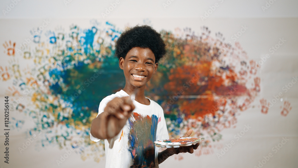 African boy standing at colorful stained wall while holding brush and point at camera. Happy highschool student wearing white shirt with stain color and look at camera with confident. Edification. African boy standing at colorful stained wall while holding brush and point at camera. Happy highschool student wearing white shirt with stain color and look at camera with confident. Edification.