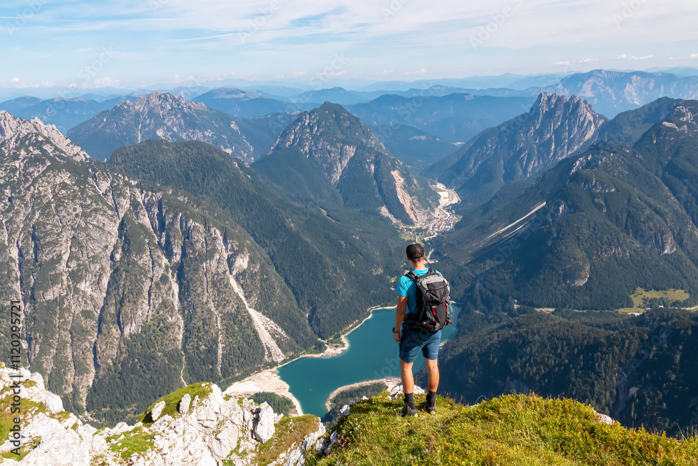 Hiker man on top of Cima Del Lago with aerial view of alpine lake Lago Del Predil in Val Canale surrounded by majestic mountain peaks of Julian Alps, Friuli Venezia Giulia, Italy. Wanderlust in nature