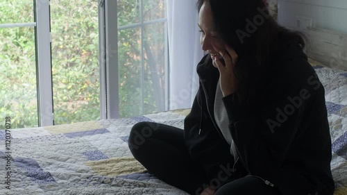Wallpaper Mural Sad woman sitting on the bed near the window, talking on the phone while looking outside. Touching her face, she reflects on feelings of loneliness and worry during the conversation Torontodigital.ca