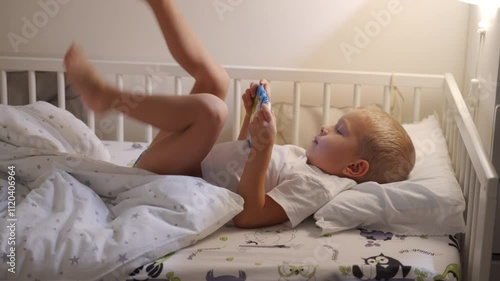 Child playing on tablet lying in bed in evening. Cute little boy watching cartoons on mobile before go to sleep. Boy enjoying in bedroom looking on screen.