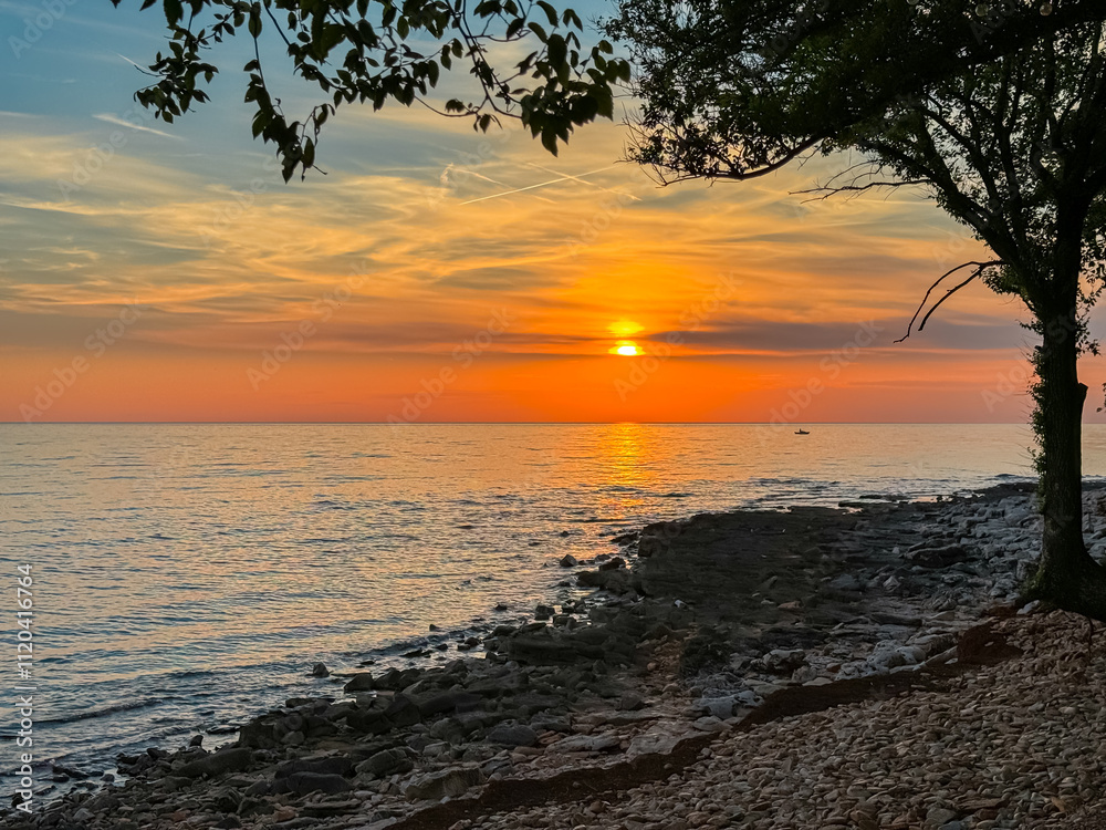 Breathtaking fiery sunset over calm Adriatic Mediterranean sea. Scenic view from coastline of Karigador, Istria peninsula, Croatia. Golden glow across sky and water. Silhouette of tree in foreground Breathtaking fiery sunset over calm Adriatic Mediterranean sea. Scenic view from coastline of Karigador, Istria peninsula, Croatia. Golden glow across sky and water. Silhouette of tree in foreground