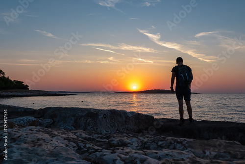 Silhouette of standing man watching breathtaking fiery sunset over calm Adriatic Mediterranean Sea in Premantura, Istria peninsula, Croatia, Europe. Summer vacation at seaside. Dramatic vibrant sky