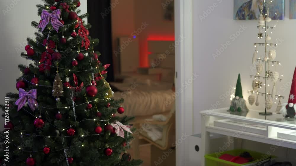 Christmas living room on Christmas Eve evening. Christmas tree is festively decorated with red balls, bows, gold ornaments and twinkling garland. Open door to bedroom.