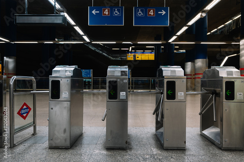 A row of turnstiles are lined up in a station. The turnstiles are silver and have a green arrow on them