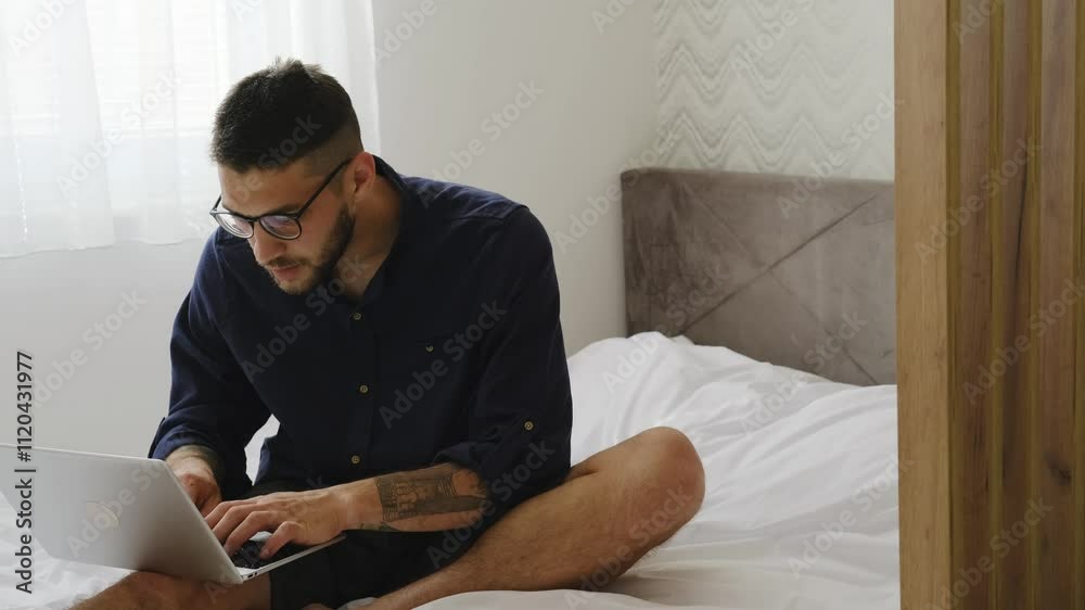 A young Caucasian bearded man sitting on a bed with a laptop. Working online from home concept. The guy is a freelancer at home interior. A young Caucasian bearded man sitting on a bed with a laptop. Working online from home concept. The guy is a freelancer at home interior.