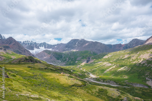 Wide green alpine valley with snake river among hills and rocks with vast view to large snow mountain top and big glacier tongue under cloudy sky. Serpentine river in high mountains. Shadows of clouds