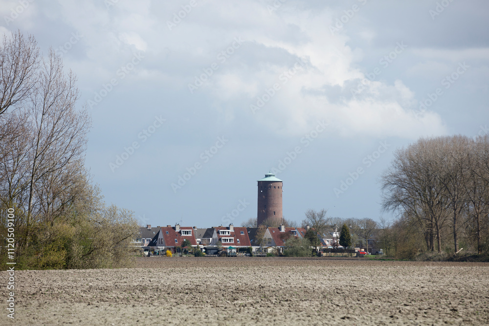 Fototapeta premium Rural Dutch landscape with houses, water tower, and plowed field. Cloudy spring day.