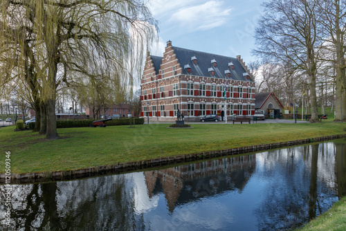 Canalside historic building, likely Dutch, with red shutters, reflected in calm water.  Mature trees and grassy areas surround the structure. photo