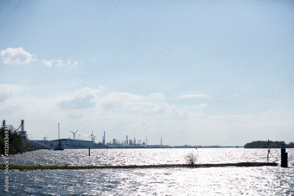 Industrial complex lines a waterway, reflecting sunlight on the rippled water.  Sunny day, light clouds.