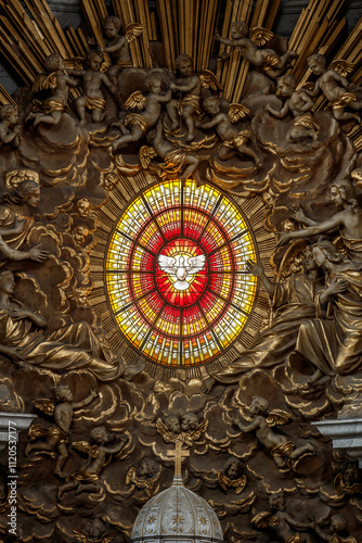 Ornate stained glass depicting the Holy Spirit, surrounded by sculpted cherubs and angels, adorns a church wall.  Religious art.