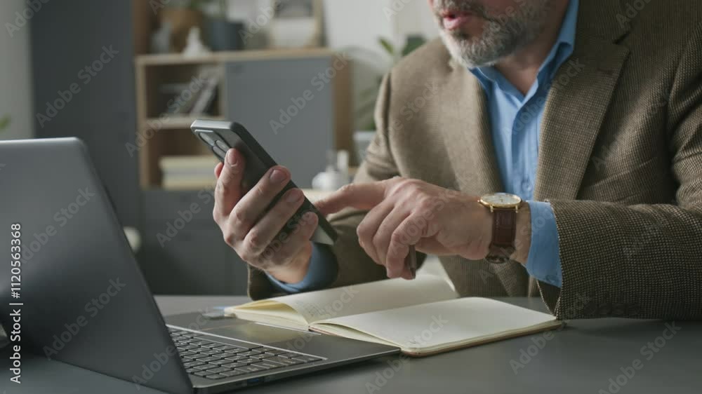 Close-up view of middle-aged businessman with grey beard sitting at desk with laptop, swiping on smartphone screen and writing down to-do list in notebook