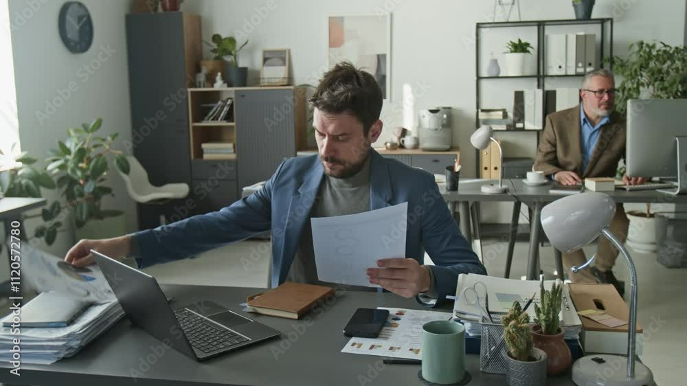 Concentrated businessman sitting at desk in open space office, reading documents and typing on laptop, then leaning back in chair and taking break during workday Concentrated businessman sitting at desk in open space office, reading documents and typing on laptop, then leaning back in chair and taking break during workday
