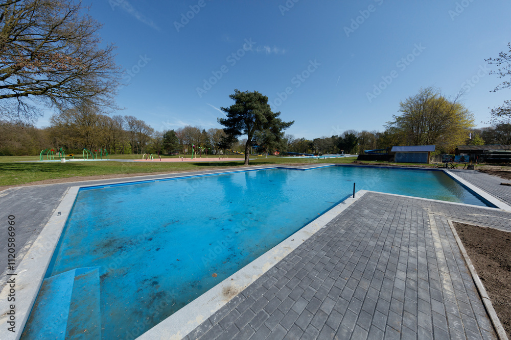 Fototapeta premium Under construction, a large blue pool sits amidst a park-like setting. Surrounding landscaping and playground equipment are visible.